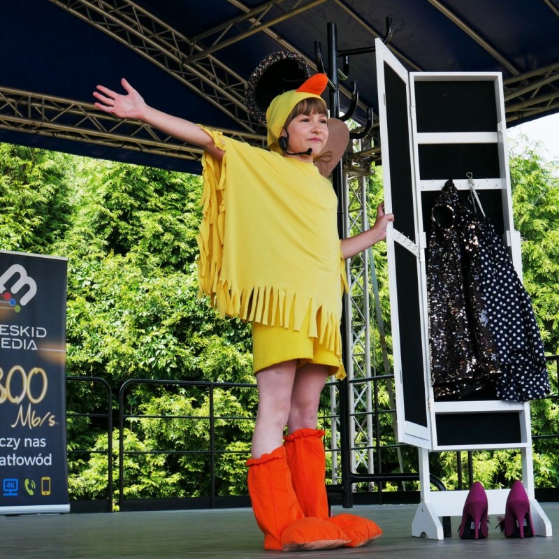 Dziewczynka przebrana za żółtego kurczaka z rozpostartymi ramionami na scenie plenerowej.