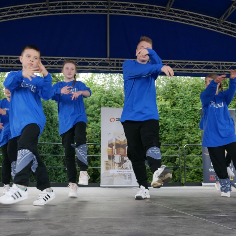 Dziecięca grupa taneczna w niebieskich bluzach podczas występu na scenie plenerowej.