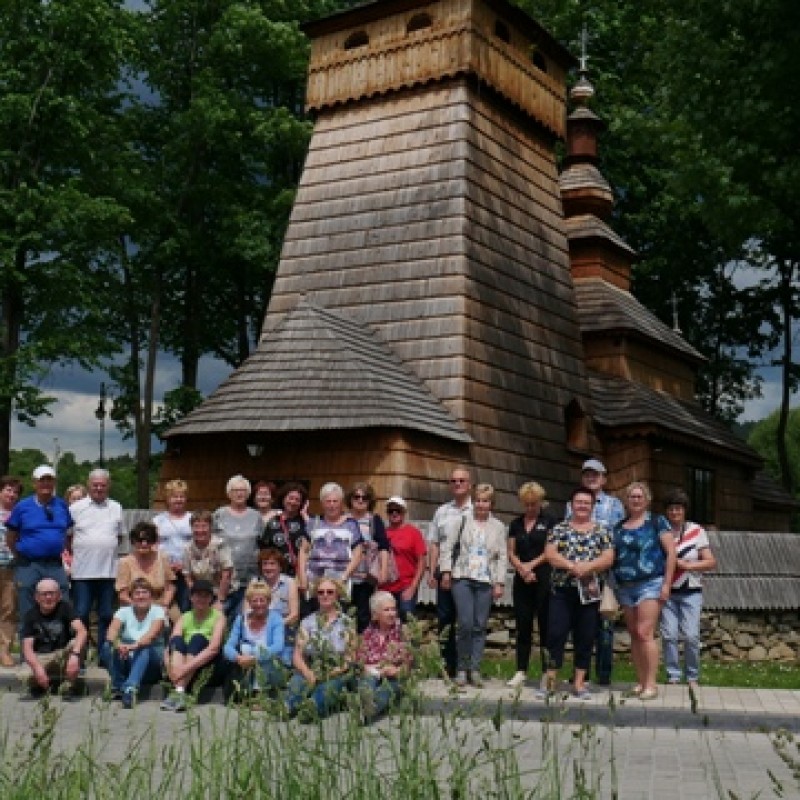 Seniorzy w grupie pozują do zdjęcia przed drewnianym kościółkiem.