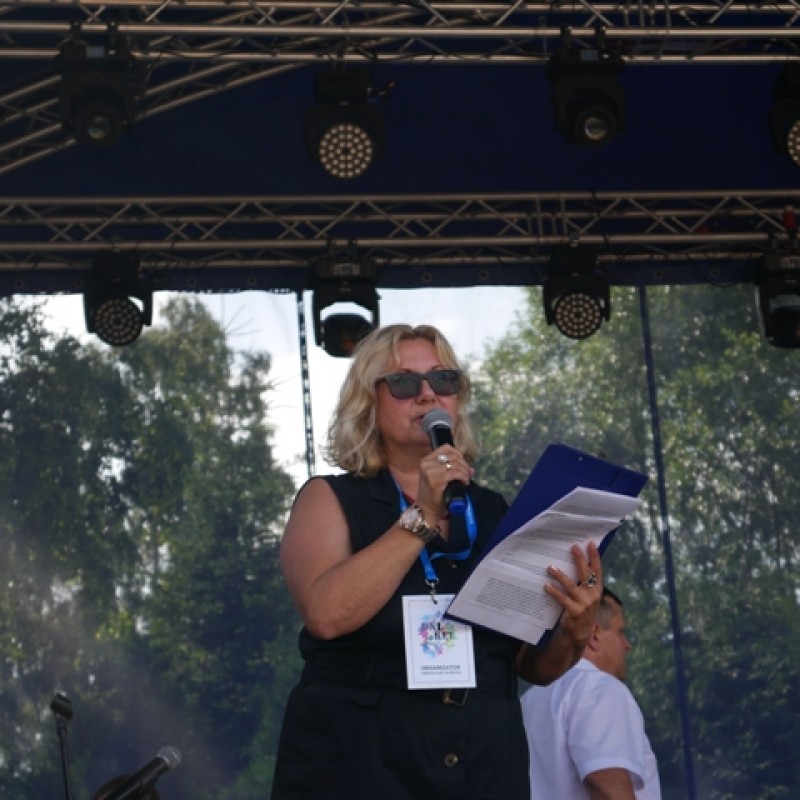 Kobieta w blond włosach i ciemnych okularach przeciwsłonecznych, stoi na scenie. W jednej ręce trzyma mikrofon, a w drugiej niebieską podkładkę.