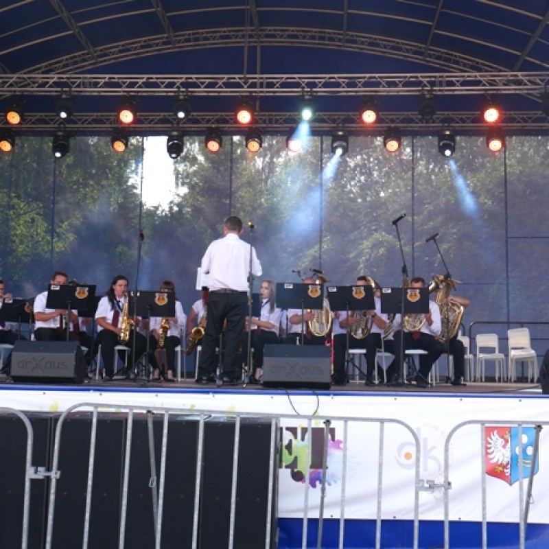 Mężczyzna w białej koszuli stoi tyłem do publiczności, dyrygując zespołowi, grających na instrumentach dętych.
