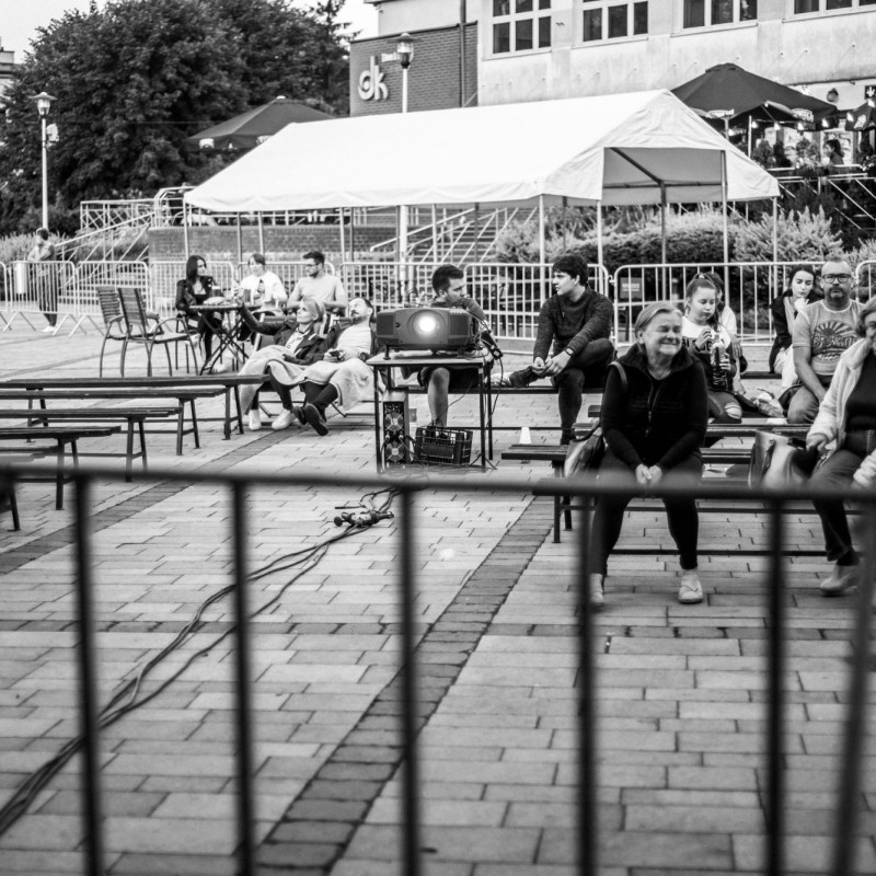 Czarno-białe zdjęcie ludzi siedzących i rozmawiających na ławkach przed sceną plenerową, za nimi stoiska.