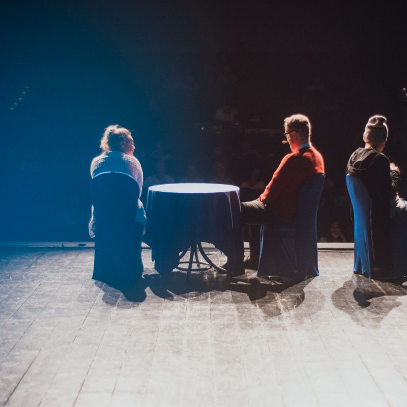 Trójka aktorów siedzi na scenie na krzesłach, dwójka przy stoliku, są zwróceni tyłem do obiektywu a przodem do publiczności.