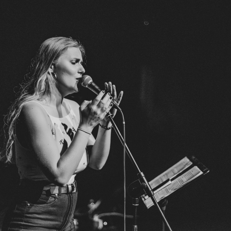 Czarno-białe zdjęcie wokalistki śpiewającej do mikrofonu, która obejmuje statyw dwoma rękami.
