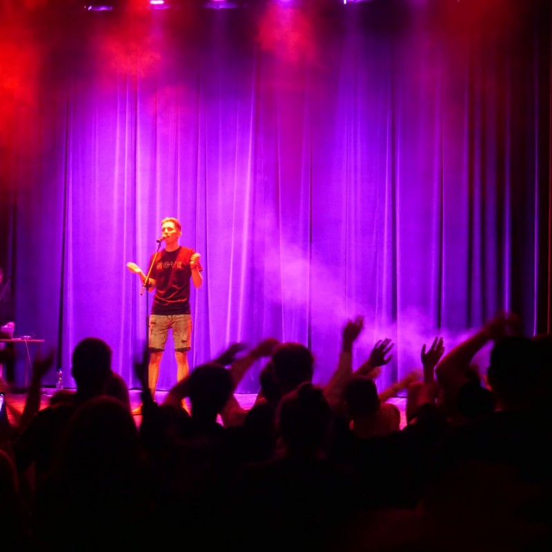 Mężczyzna mówi do mikrofonu na statywie na scenie widowiskowej w oświetleniu fioletowych i czerwonych reflektorów, przed nim publiczność.