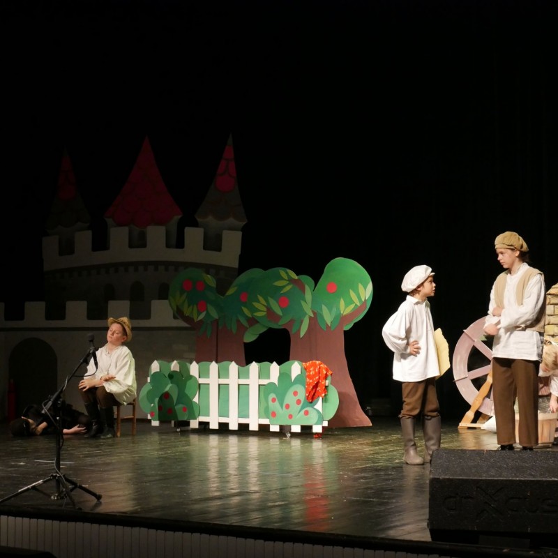 Kilkoro młodych aktorów podczas spektaklu w wiejskiej scenerii na scenie widowiskowej. 