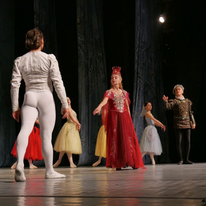 Baletmistrz zwrócony w stronę baleriny w czerwonej długiej sukni i koronie, w tle inni tancerze.
