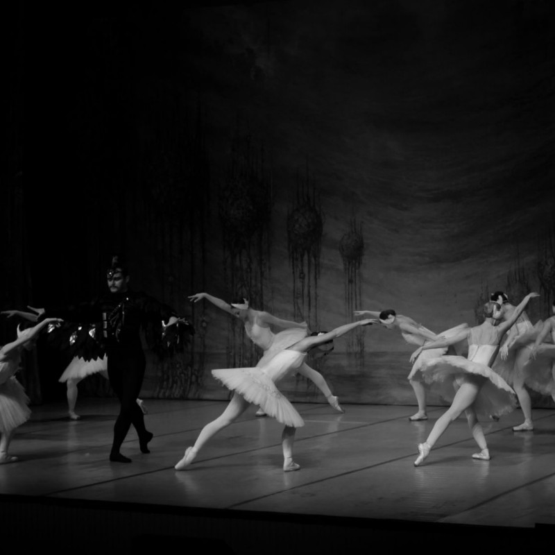 Czarno-białe zdjęcie, na przedzie ubrany na czarno tancerz, a na około niego tancerki w skłonie z jedną ręką wyciągniętą do przodu.