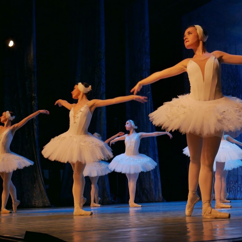 Tancerki ubrane w białe sukienki ze skrzyżowanymi nogami i rękoma rozpostartymi na boki.