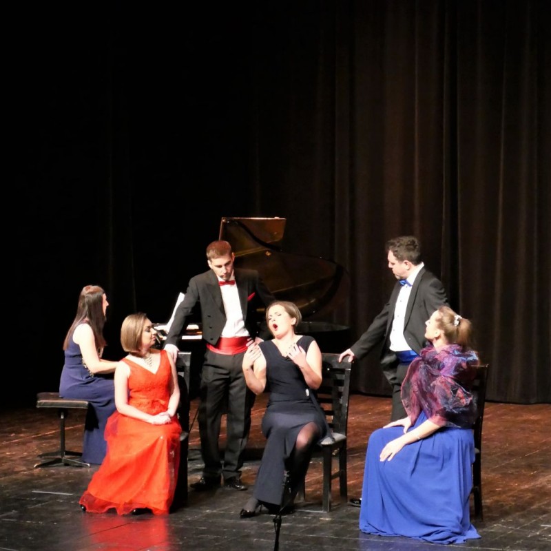 Na scenie sześciu muzyków. Jedna kobieta gra na fortepianie. Trzy kobiety siedzą na krzesłach śpiewając. Dwoje mężczyzn stoi przy nich. 