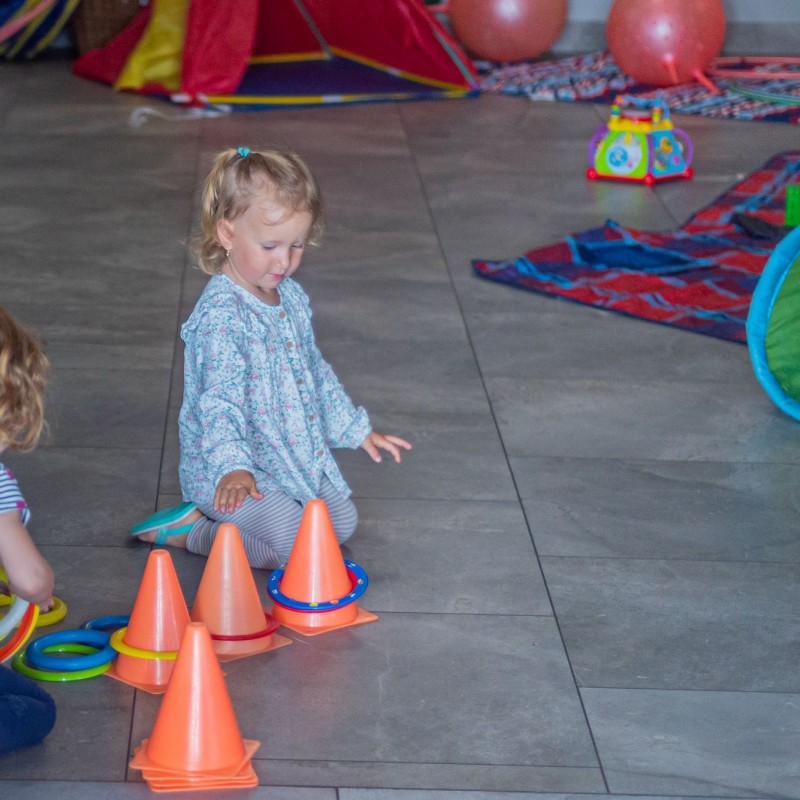 Fot. Łukasz Kuc/Dwie dziewczynki bawią się obręczami narzucając je na pachołki na ziemi, w tle zabawki oraz materiałowe przedmioty do toru przeszkód.