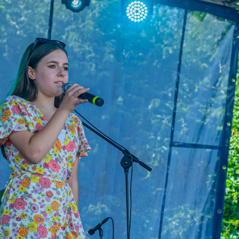Fot. Łukasz Kuc/Dziewczyna z sukience w kwiaty śpiewa do mikrofonu na scenie plenerowej domu kultury.