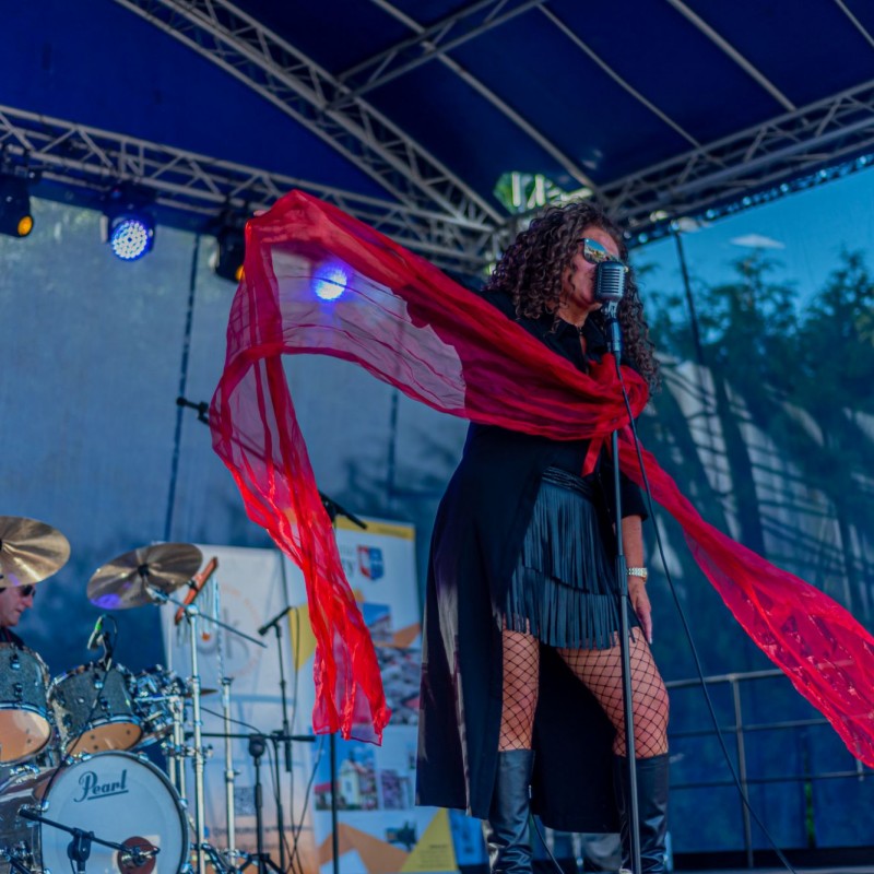 Fot. Łukasz Kuc/Kobieta stoi przy mikrofonie na scenie plenerowej, wyrzuca ręką czerwoną chustę do boku. Za nią perkusista.