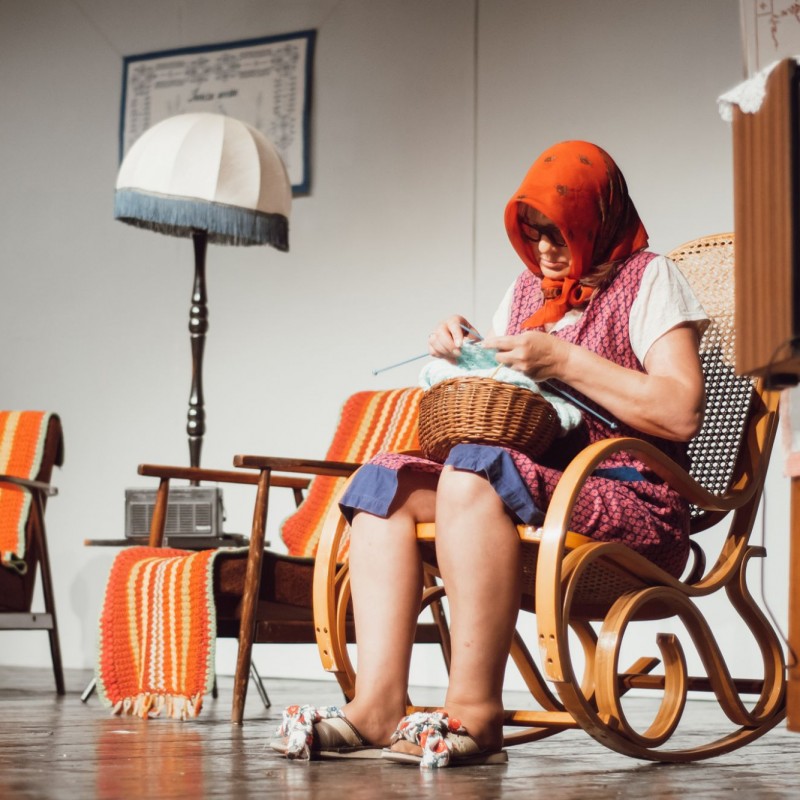 Fot. Łukasz Kuc/Scena domu kultury, teatralna dekoracja wnętrza domu wśród której siedzi kobieta stylizowana na starszą panią na fotelu bujanym szydełkując. 