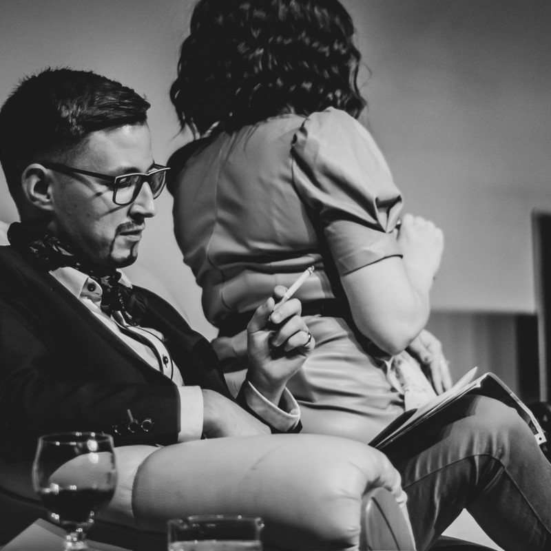 Fot. Łukasz Kuc/Czarno-białe zdjęcie, mężczyzna siedzi na fotelu i trzyma papierosa w ręce, kobieta siedzi na oparciu fotela zwrócona do niego tyłem.