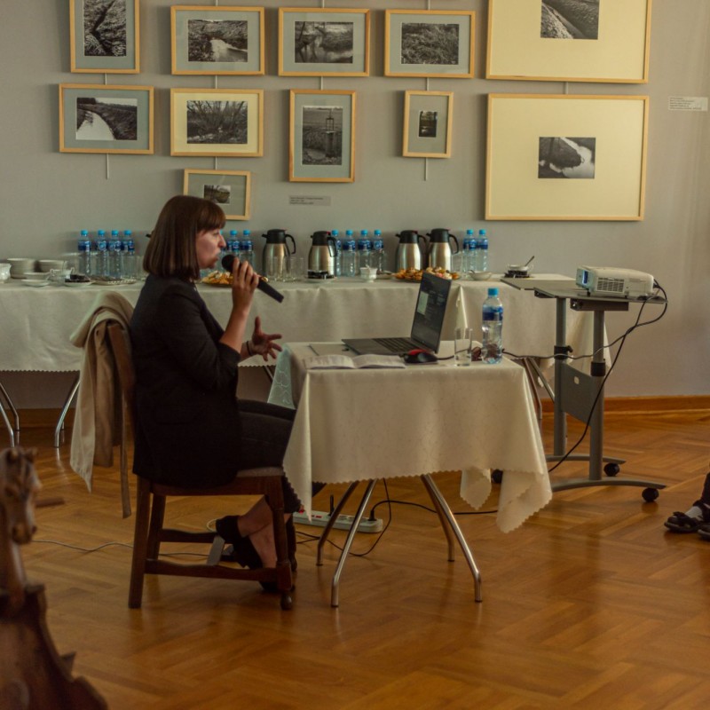 Fot. Łukasz Kuc/Kobieta siedzi przy stoliku z laptopem trzymając mikrofon przy ustach, przed nią grupa ludzi, a obok miejsce na wodę i gorące napoje.