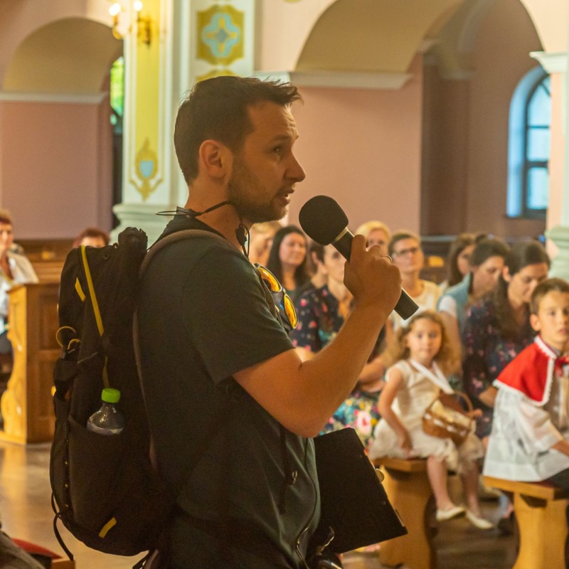 Fot. Łukasz Kuc/W kościele siedzą ludzie przed nimi mężczyzna z mikrofonem i plecakiem.