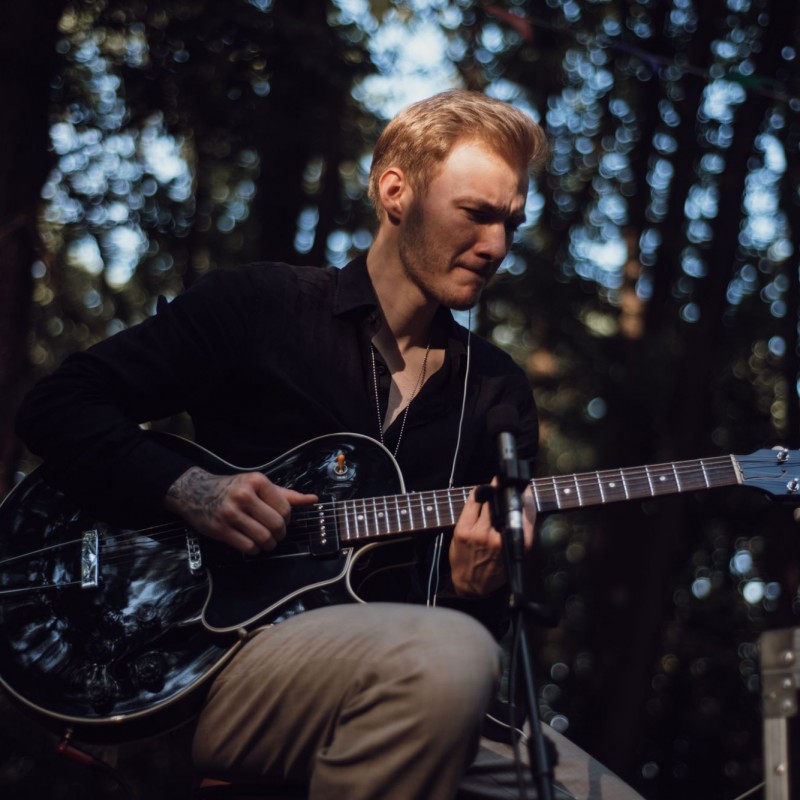 Fot. Łukasz Kuc/Mężczyzna gra na czarnej gitarze, w tle las.