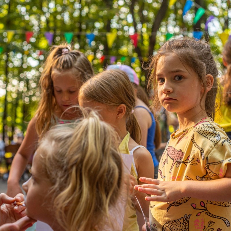Fot. Łukasz Kuc/Dzieci ustawione w kolejce do malowania twarzy, w tle las i zawieszone girlandy.