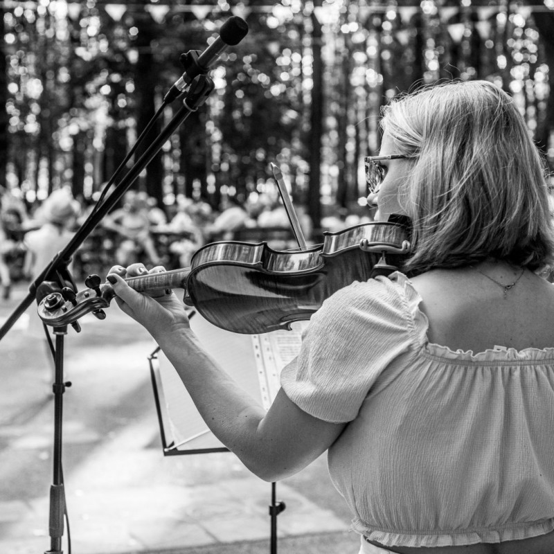 Fot. Łukasz Kuc/Dziewczyna odwrócona plecami do aparatu gra na skrzypcach, przed nią mikrofon, dalej słuchacze.