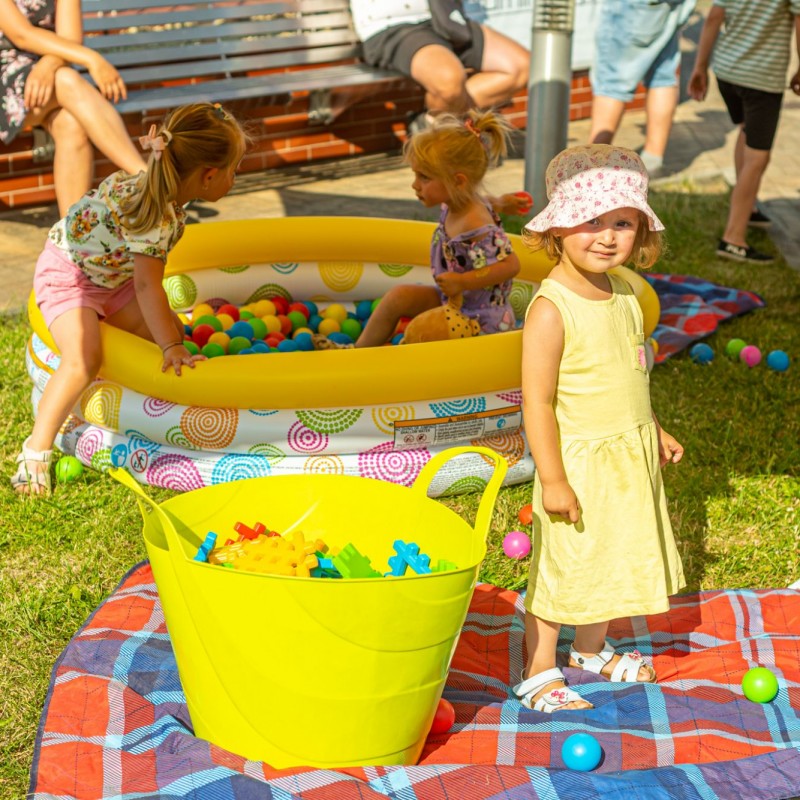 Fot. Łukasz Kuc/Dzieci bawią się w basenie z kolorowymi kulkami, przed nimi dziewczynka stoi na kocu obok zabawek i patrzy w kamerę.