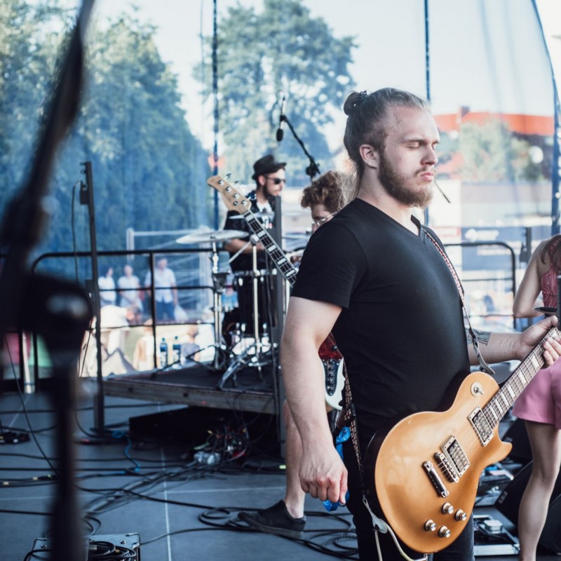 Fot. Łukasz Kuc/Gitarzysta z gitara elektryczna w dynamicznej pozie podczas występu, w tle reszta zespołu.