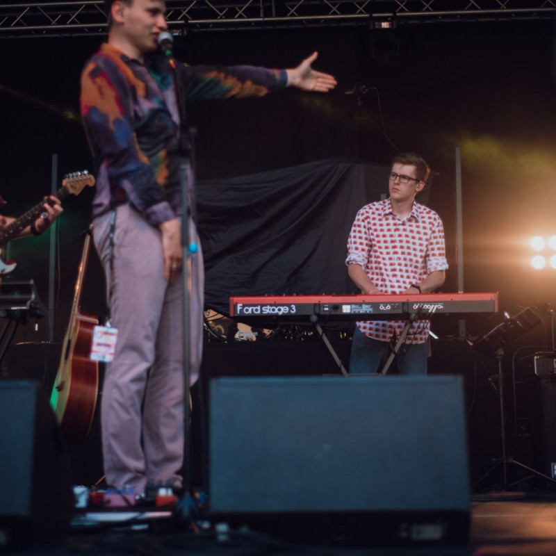 Fot. Łukasz Kuc/Wokalista zespołu stojący przy mikrofonie wskazuje ręka na innego członka zespołu, w tle pianista grający na keyboardzie.